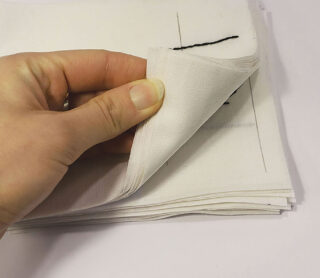 Eine Hand hebt das oberste Blatt eines Stapels weißer Stoffquadrate auf einer weißen Oberfläche an.