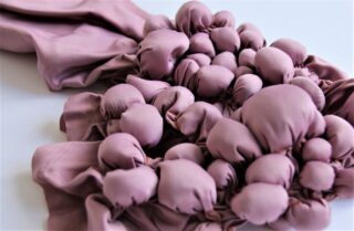 Eine Nahaufnahme von malvenfarbenem Stoff, der sich vor einem schlichten Hintergrund zu zahlreichen runden, bauschigen Büscheln zusammenballt.