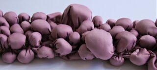 Mehrere Büschel malvenfarbenen Stoffs sind vor einem schlichten hellen Hintergrund gerafft und zu runden, bauchigen Formen zusammengebunden.
