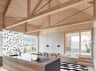 Moderne Küche und Essbereich mit hellen Holzoberflächen, großen Fenstern, schwarz-weißen Akzenten und freiliegenden Deckenbalken. Vorhang und Teppich mit Kuhfellmuster setzen dekorative Akzente.