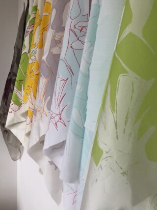 Vor einem weißen Hintergrund hängen in einer Reihe mehrere halbtransparente Stoffbahnen in unterschiedlichen Farben, jeweils mit einem Blumen- oder Blättermuster.