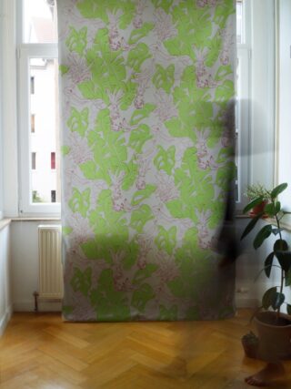 Ein mit einem Vorhang mit grünem Blättermuster bedecktes Fenster in einem hellen Raum mit Parkettboden und einer Topfpflanze in der Nähe.