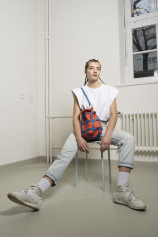 Eine Frau mit geflochtenen Haaren sitzt auf einem Stuhl in einem minimalistischen Raum. Sie trägt ein weißes T-Shirt, helle Jeans, weiße Turnschuhe und hält eine rot-blaue Tasche in der Hand.