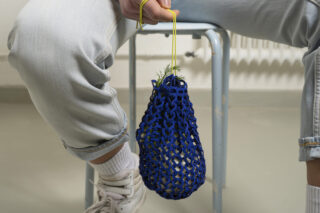 Eine Person sitzt auf einem Metallhocker und hält einen blauen Netzbeutel mit gelben Griffen, in dem sich grüne Blätter befinden. Die Person trägt helle Jeans, weiße Socken und weiße Schuhe.