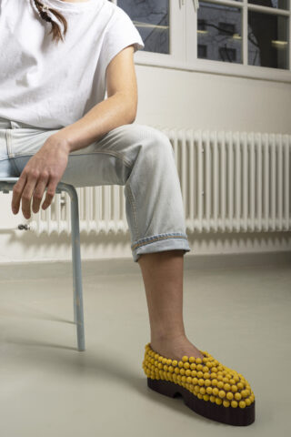 Person, die auf einem Stuhl sitzt und hellblaue Jeans, ein weißes T-Shirt und gelbe Schuhe mit einer strukturierten, kugelartigen Oberfläche trägt.
