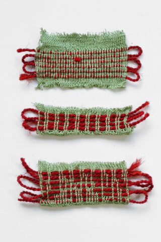 Drei kleine rechteckige gewebte Stoffmuster mit grünen und roten Fäden, vertikal angeordnet auf einem weißen Hintergrund.