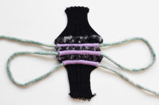 Ein gestricktes schwarzes und mehrfarbiges Objekt mit vier grünen Filzsträngen, die von jeder Seite ausgehen, auf einem weißen Hintergrund.