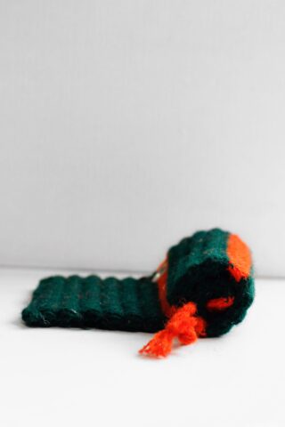 Ein aufgerollter, gerippter grüner Stoff mit einem orangefarbenen Streifen und losen orangefarbenen Fäden auf einer weißen Oberfläche vor einem einfarbigen Hintergrund.