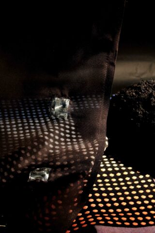 Nahaufnahme eines schwarzen strukturierten Stoffes mit Metallverzierungen und einem Netzabschnitt, der durch gerichtetes Licht ein gepunktetes Schattenmuster erzeugt.