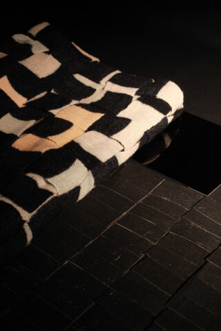 Ein gefalteter schwarz-weißer Textilstoff mit geometrischem Muster liegt auf einer Oberfläche, die mit glänzenden schwarzen rechteckigen Fliesen bedeckt ist.