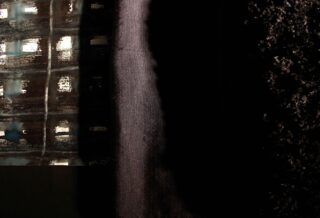 Eine dunkle, strukturierte Oberfläche nimmt die rechte Seite ein, während die linke Seite ein Raster aus beleuchteten Fenstern zeigt, was an einen Teil eines Gebäudes bei Nacht erinnert.