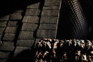 Eine Nahaufnahme strukturierter Oberflächen, darunter gitterartige schwarze Kacheln, eine reflektierende schwarze Ebene und eine Reihe komprimierter, zerknitterter Materialien mit gemischten Farben unter dramatischen Schatten.