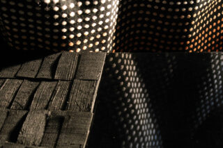 Nahaufnahme eines gemusterten Stoffes mit weißen Punkten, dessen Spiegelbild auf einer glänzenden schwarzen Oberfläche neben einem strukturierten, gewebten schwarzen Material sichtbar ist.