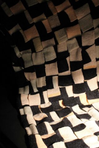 Eine Nahaufnahme eines gewebten Textils mit einem Schachbrettmuster aus schwarzen, weißen und beigen Quadraten, das eine strukturierte, dreidimensionale Oberfläche erzeugt.