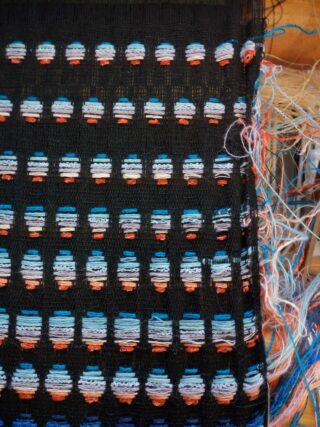 Ein gewebter Stoff mit einem Raster aus ovalen Lücken, bestehend aus horizontalen Fäden in Blau, Weiß, Orange und Schwarz und losen Fäden an der Seite.