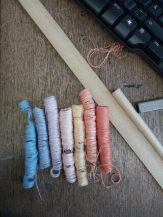 Sieben Garnrollen in verschiedenen Pastellfarben sind auf einer Holzoberfläche neben einem Lineal, einer Tastatur und einem kleinen rechteckigen Objekt angeordnet.