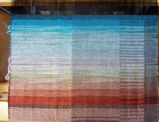 Ein auf einem Webstuhl gewebter Textilstoff weist horizontale Abstufungen von Blau-, Grau- und Rotbrauntönen mit sichtbaren Kett- und Schussfäden auf.