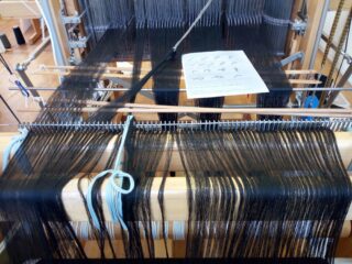 Ein Webstuhl mit aufgespannten schwarzen Fäden und einem darauf platzierten Anleitungsblatt, aus der Perspektive des Bedieners betrachtet.