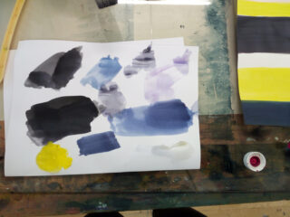 Ein Blatt Papier mit Flecken aus schwarzer, blauer, gelber und violetter Wasserfarbe liegt auf einem unordentlichen Tisch neben einem Gemälde mit schwarzen, gelben und weißen Streifen.