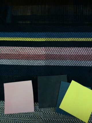 Drei farbige Quadrate – Rosa, Schwarz und Gelb – sind auf einem strukturierten Stoff mit horizontalen Streifen in Blau, Gelb, Rosa und Schwarz platziert.