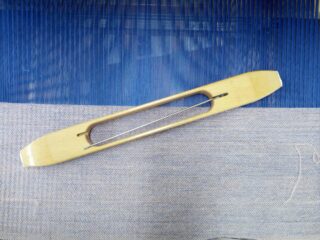 Ein hölzerner Weberschiffchen liegt auf blauem und beigem Stoff, darunter sind Fäden auf einem Webstuhl sichtbar.