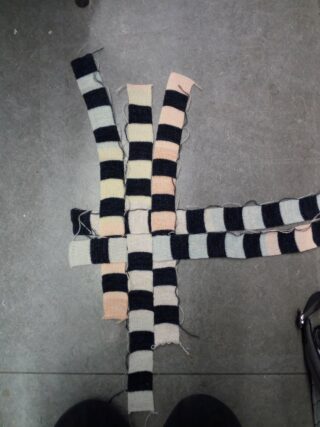 Vier Streifen aus gestreiftem Strickstoff, abwechselnd in hellen und dunklen Farben, sind miteinander verwoben und auf einem grauen Boden ausgelegt.