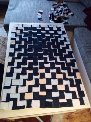 Auf einer Tischplatte ist ein teilweise zusammengesetztes Patchwork aus schwarzen, beigen und hellrosa gestrickten Quadraten und Rechtecken zu sehen; weitere Teile liegen auf einem Sofa in der Nähe.