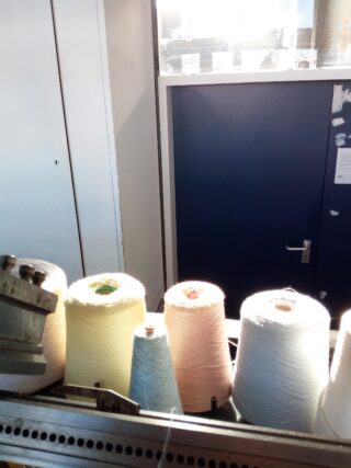 Mehrere große Spulen mit pastellfarbenem Garn sind auf einem Metallgestell in der Nähe einer blauen Tür in einem hell erleuchteten Raum angeordnet.