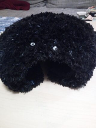 Ein flauschiges, schwarzes Nackenkissen in U-Form mit zwei kleinen Wackelaugen auf einer hellen Oberfläche.