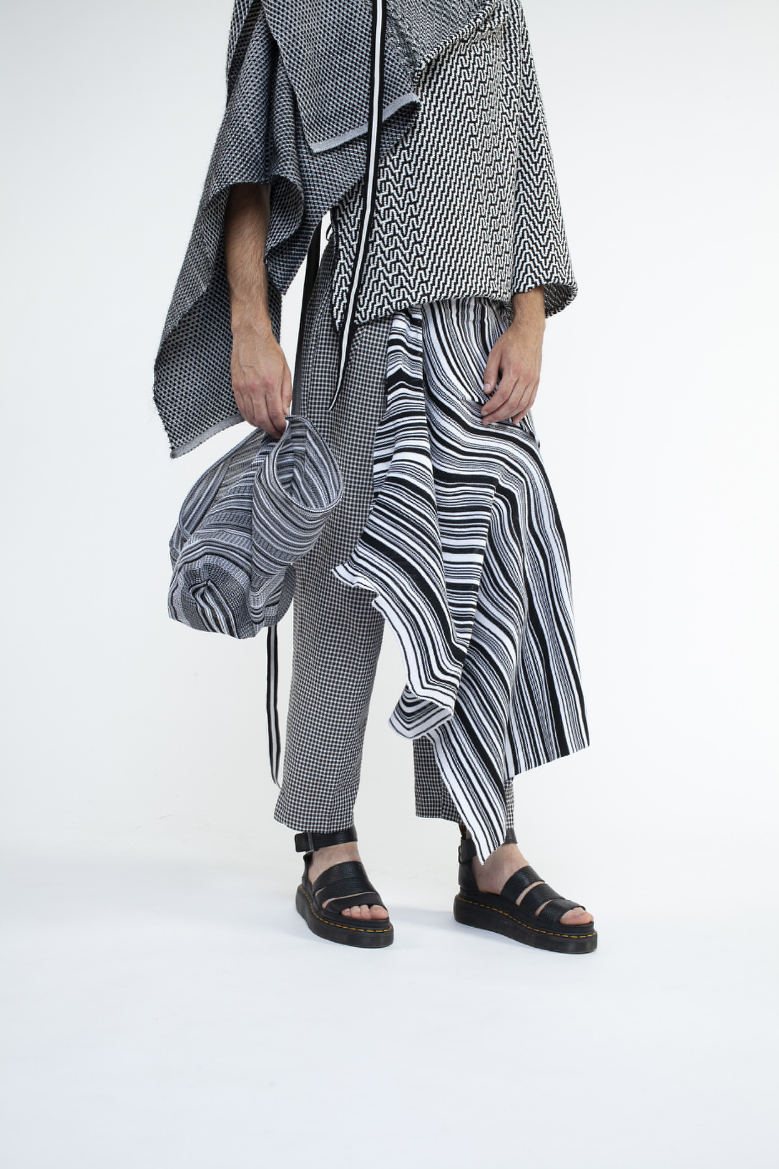 Person in schwarz-weiß gemusterter Kleidung und Sandalen, hält eine passende Tasche und steht vor einem schlichten hellen Hintergrund.