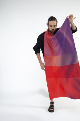 Eine Person mit langen Haaren und Bart hält einen großen, rot-violetten Farbverlaufsstoff vor einem schlichten weißen Hintergrund hoch.