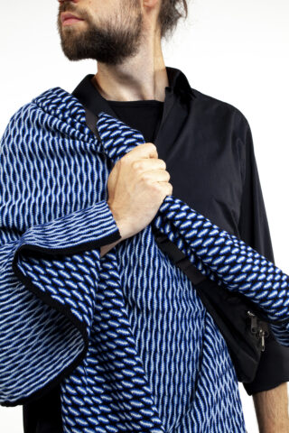 Eine Person mit schwarzem Hemd hält einen blau-weiß gemusterten Stoff über der Schulter, der Kopf ist leicht zur Seite gedreht.