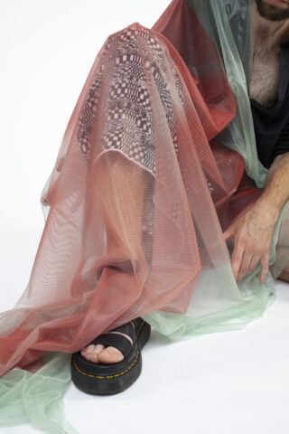 Eine Person, die gemusterte Leggings und schwarze Sandalen trägt, ist teilweise mit durchsichtigem roten und grünen Stoff bedeckt und sitzt vor einem schlichten Hintergrund.