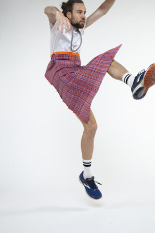 Eine Person, die ein weißes Hemd, einen gestreiften Rock, gestreifte Socken und Turnschuhe trägt, ist mitten im Sprung vor einem schlichten weißen Hintergrund zu sehen.