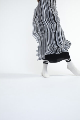 Person, die einen fließenden schwarz-weiß gestreiften Rock, eine schwarze Hose und weiße Socken mit schwarzen Streifen trägt und vor einem schlichten weißen Hintergrund posiert.