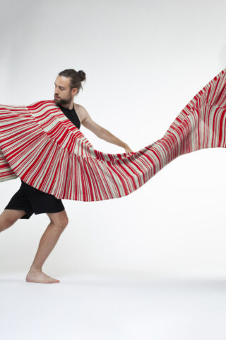 Eine barfüßige Person in schwarzer Kleidung hält und breitet einen großen, plissierten rot-weiß gestreiften Stoff vor einem schlichten weißen Hintergrund aus.