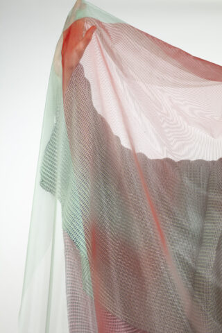 Eine Person steht hinter mehrschichtigen, durchscheinenden Stoffen in Rot-, Grün- und Grautönen, die ihre Form vor einem schlichten Hintergrund teilweise verdecken.
