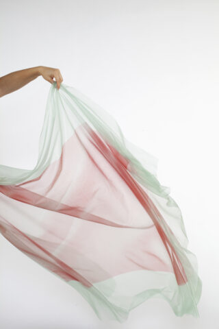 Eine Hand hält einen durchscheinenden, hellgrünen und rosa Stoff vor einem schlichten weißen Hintergrund hoch.