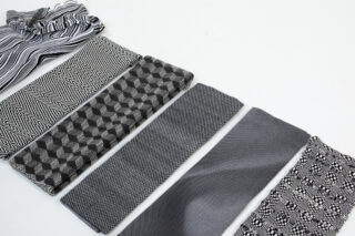 Sechs gefaltete Stoffstücke mit geometrischen oder gemusterten Mustern in Schwarz, Weiß und Grau sind in einer Reihe auf einer weißen Oberfläche angeordnet.