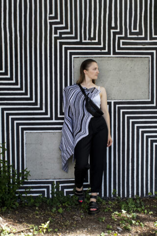 Eine Frau steht vor einer geometrisch schwarz-weiß gestreiften Wand. Sie trägt schwarze Hosen, Sandalen, eine Umhängetasche und ein asymmetrisches Oberteil mit passenden Streifen.