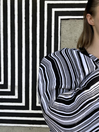 Eine Person in schwarz-weiß gestreiftem Kleidungsstück steht vor einer Wand mit einem geometrischen schwarz-weißen Streifenmuster. Nur ein Teil ihres Gesichts und Oberkörpers ist sichtbar.