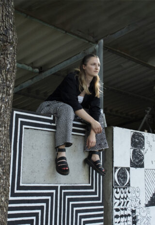 Eine Frau sitzt auf einem mit schwarz-weißen geometrischen Mustern verzierten Betonvorsprung unter einem Metalldach und blickt in die Ferne.