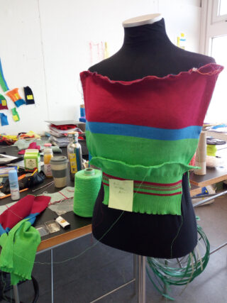 Eine Schneiderpuppe zeigt ein halbfertiges Strickstück in Rot, Blau und Grün. Im Hintergrund sind Garnkegel, Werkzeuge und Bastelbedarf auf einem Tisch verstreut.