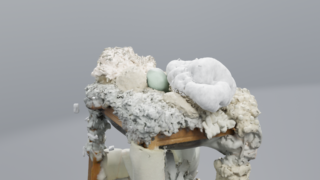 Ein kleiner Holztisch ist mit einem unregelmäßig strukturierten, weiß-grauen, schaumstoffartigen Material bedeckt. Darauf liegen eine hellblaue Kugel und eine weiche weiße Form. Der Hintergrund ist schlicht grau.