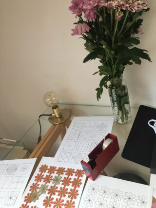 Ein Arbeitsplatz mit geometrischen und floralen Musterskizzen, einem roten Klebebandspender, einer Lampe und einer Vase mit lila Blumen auf einem Glasschreibtisch.