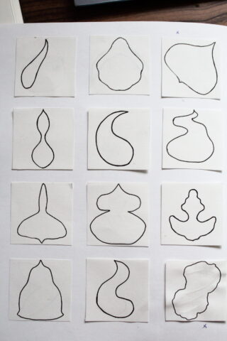 Zwölf unregelmäßige, abstrakte Formen, die auf einzelnen weißen Quadraten umrissen sind, sind in einem 3x4-Raster auf einem Blatt Papier angeordnet.