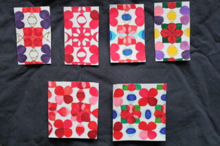 Sechs kleine rechteckige Papiere, jedes mit unterschiedlichen bunten geometrischen und floralen Mustern, sind in zwei Reihen auf einem dunklen Stoffhintergrund angeordnet.