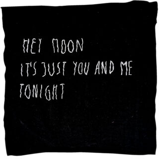 Auf einem schwarzen Stoffquadrat ist in unregelmäßigen Buchstaben der weiß gestickte Text „HEY MOON, IT’S JUST YOU AND ME TONIGHT“ aufgedruckt.