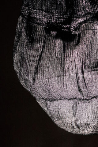 Eine Nahaufnahme eines strukturierten, halbtransparenten Stoffobjekts, das von hinten beleuchtet wird, wodurch sichtbare Falten und Schatten entstehen.