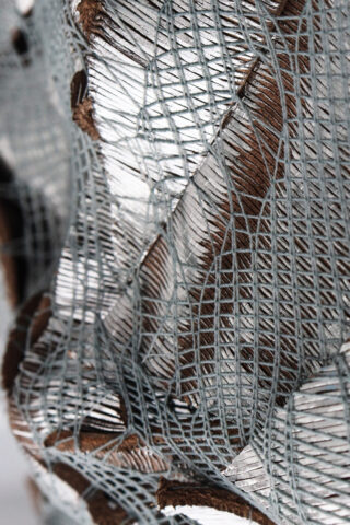 Nahaufnahme einer strukturierten Oberfläche mit sich kreuzenden silbergrauen Fäden und braunen Bereichen, die ein komplexes, netzartiges Muster mit einigen gebrochenen oder zerrissenen Abschnitten zeigt.